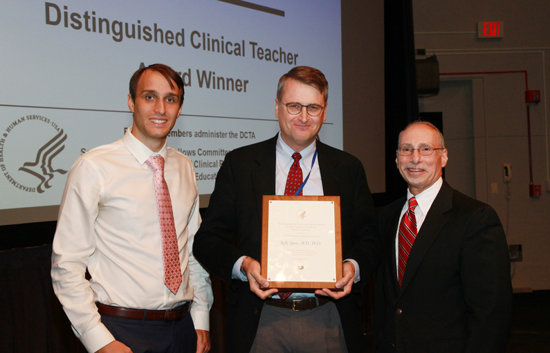 Dr. Christopher Pleyer, Dr. Kelly Stone and Dr. Robert Lembo stand in front of a screen that says Distinguished Clinical Teacher Award Winner. Stone holds a plaque