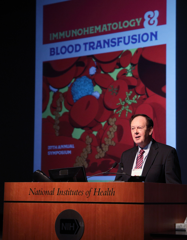 Dr. William Ward speaks at a podium and a screen behind him is a poster that says Immunohematology & Blood Transfusion, 27th Annual Symposium