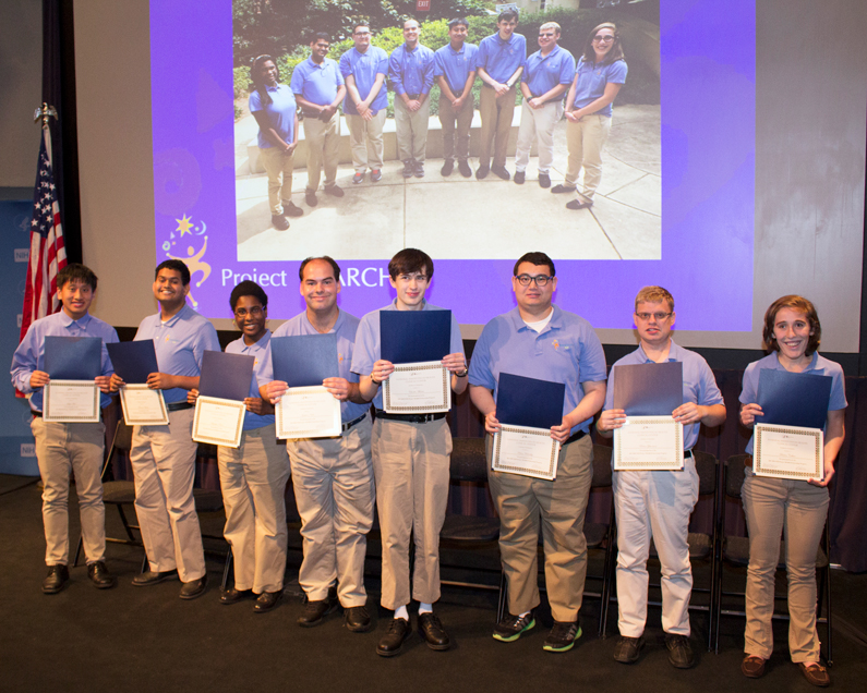 Eight young men and women line up holding graduation certificates in Lipsett Auditorium